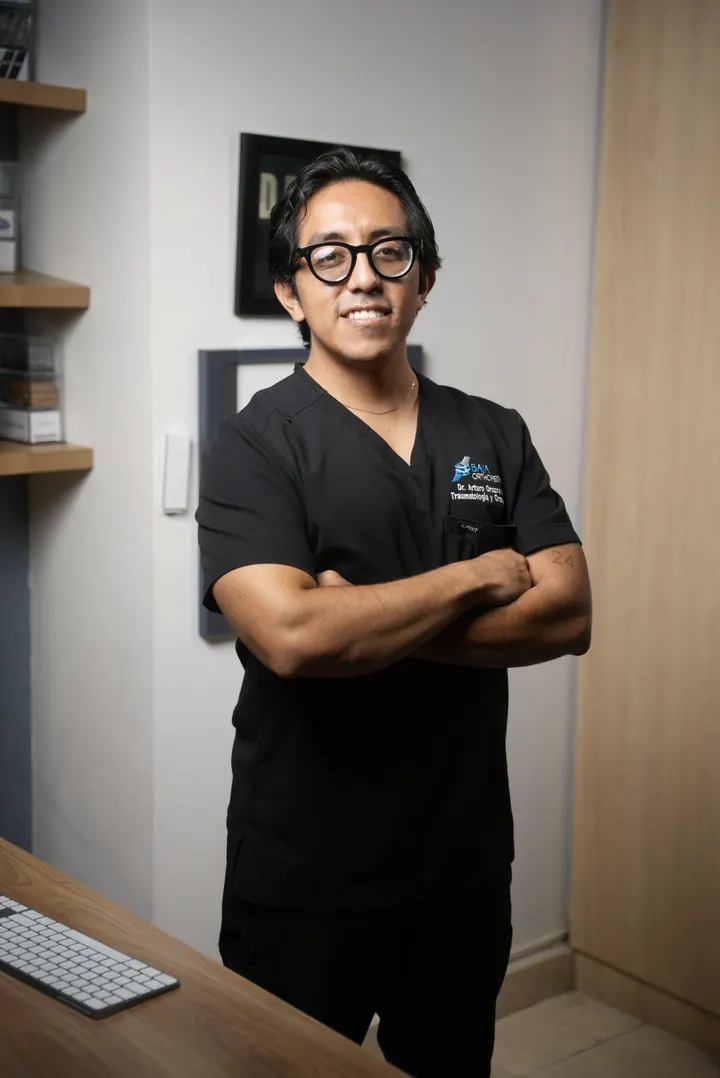 El Dr. Arturo Orozco de pie en su consultorio, con uniforme quirúrgico negro y los brazos cruzados